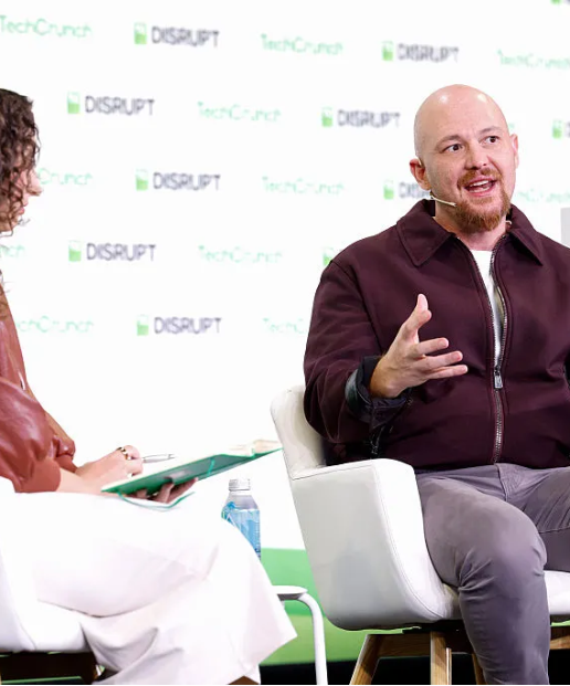 TechCrunch DISRUPT 로고가 반복 인쇄된 흰색 배경 앞 흰색 의자에 자주색 재킷 차림의 남성이 앉아 한 손을 들어 말하고, 화면 왼쪽 끝에 초록색 서류철을 든 사람이 일부 보이는 사진.